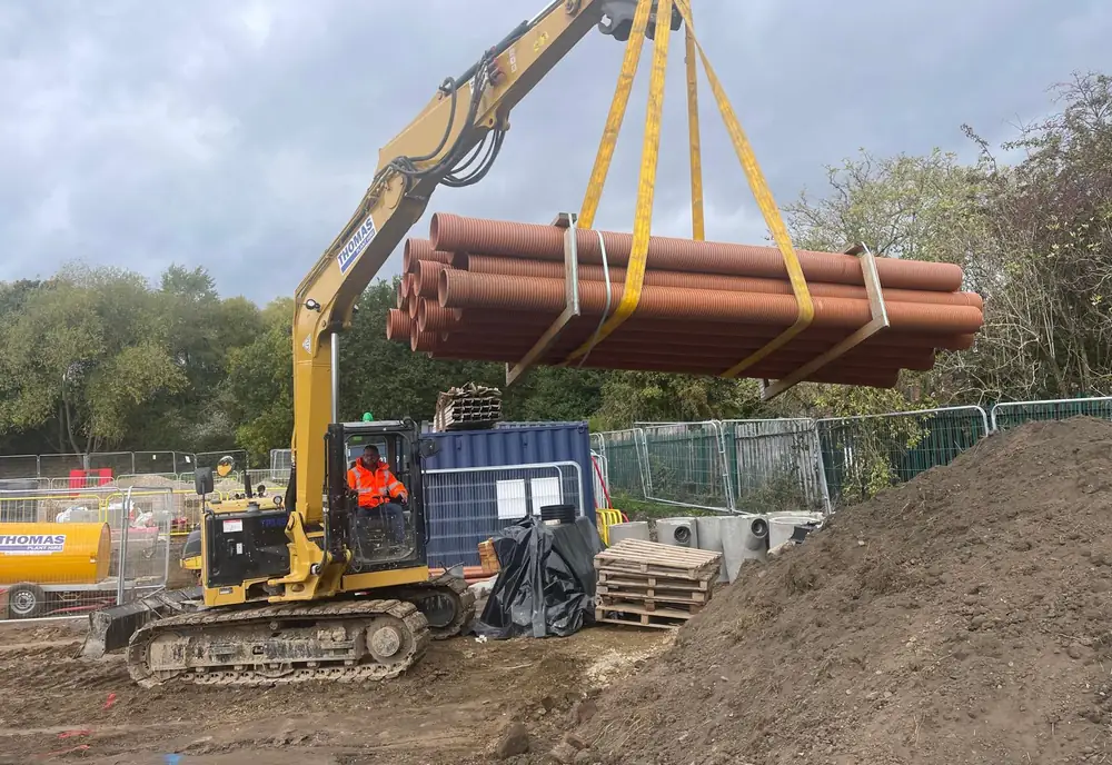 A tracked 360-degree excavator lifting a banded bundle of drainage pipes on slings from a spreader beam