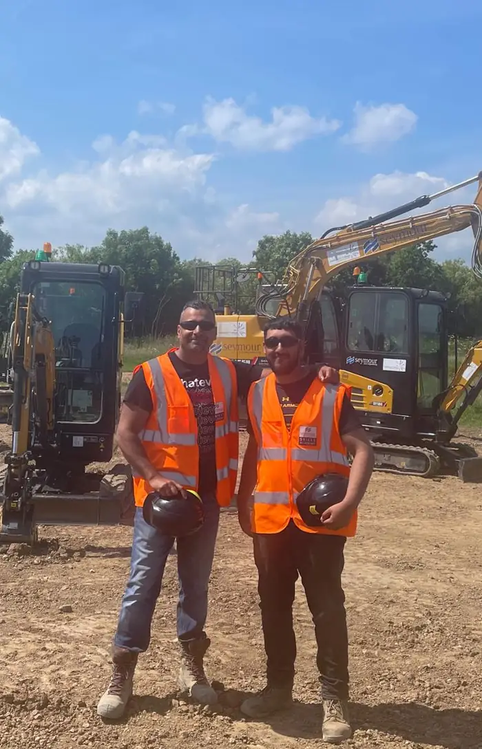 Two people in hi-vis and hard hats standing in front of a tracked mini excavator on a site