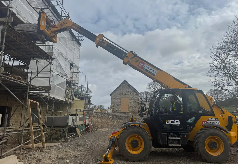 A JCB telescopic handler with its boom extended alongside a scaffolded stone building